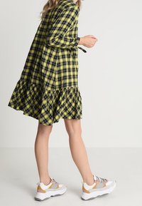 Abito a quadri giallo e nero con maniche lunghe e orlo arricciato, abbinato a sneakers bianche, grigie e arancioni. Tessuto dalla consistenza liscia.