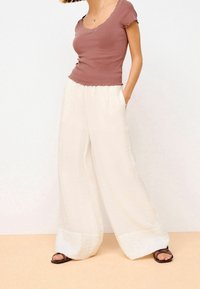 Femme portant un top ajusté rose poussiéreux à manches courtes et un pantalon blanc large et ample, les mains dans les poches et des sandales sombres.