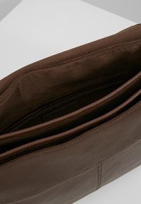 Hnědá kožená taška s viditelným šitím, otevřená a ukazující tmavě látkový interiér a vnitřní přihrádky.