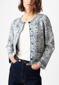 Mėlynos ir baltos spalvos raštų švarkas, kuriame yra apvali iškirptė, priekiniai mygtukai ir tekstūruota medžiaga su geometriniais raštais. Pora su balta palaidine.