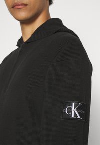 Czarna bluza z kapturem, wykonana z teksturowanej tkaniny, z zapinanym przodem, charakteryzująca się płaskim kapturem z dzianiny oraz boczną naszywką z logo "Calvin Klein Jeans".