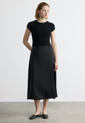 Μια γυναίκα στέκεται μπροστά σε λευκό τοίχο, φοράει μαύρο πλεκτό τοπ με κοντά μανίκια, μαύρη μεσαίου μήκους φούστα και μαύρα σανδάλια με λουράκι στο πίσω μέρος.