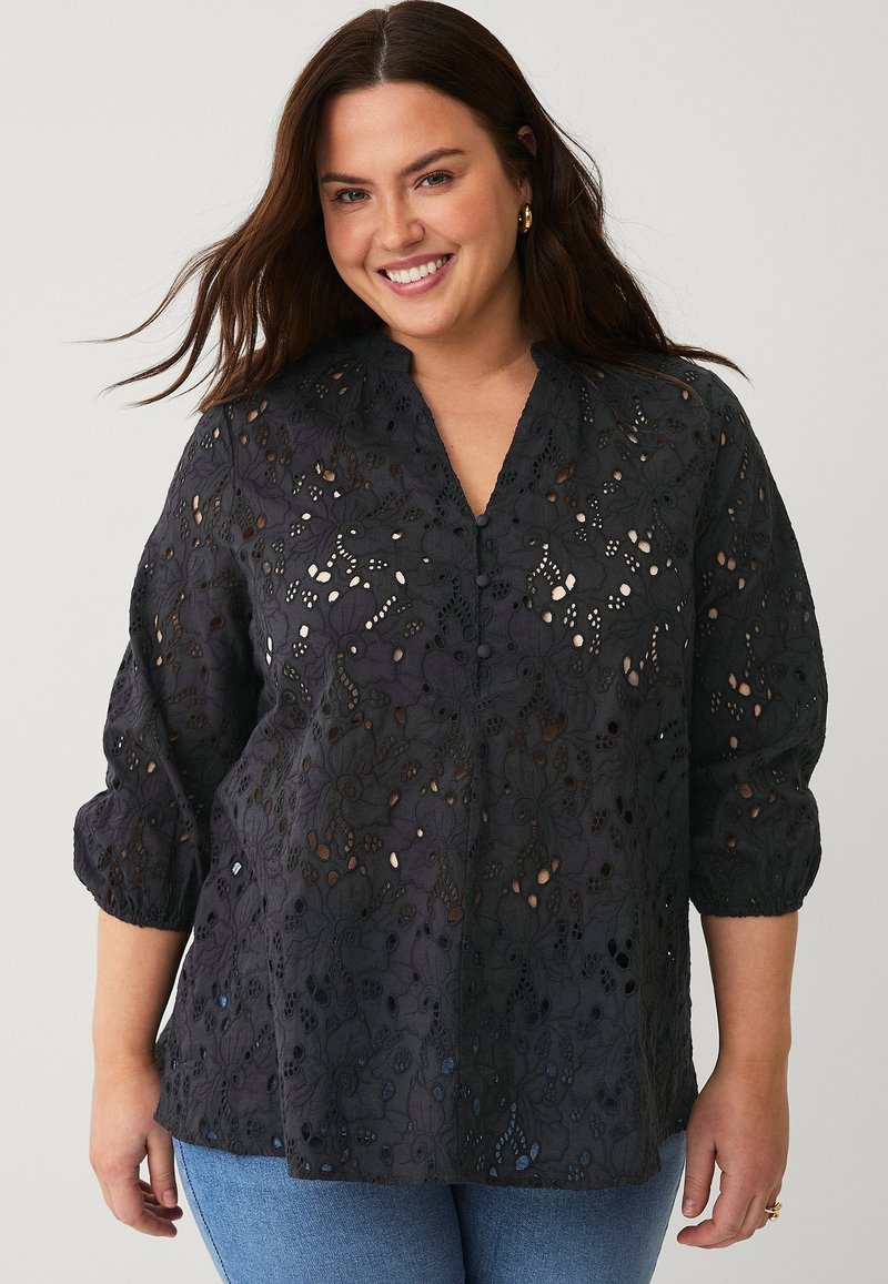 Blusa nera ricamata con intricate aperture floreali, maniche a 3/4, scollatura a V e dettagli di bottoni sottili; abbinata a jeans blu.