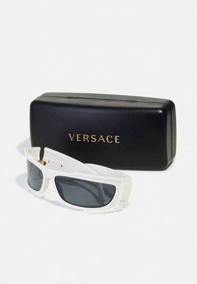Hvide oversized solbriller med mørke linser, der har et dristigt design og guldhardware, placeret ved siden af et sort Versace-etui.