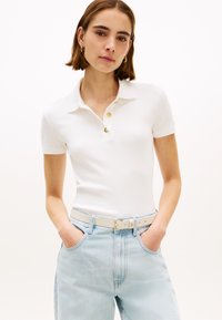 Polo blanc à manches courtes avec col, trois boutons dorés et une coupe ajustée. Associé à un jean denim bleu clair et une ceinture blanche.