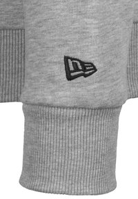 Puño de sudadera gris con textura acanalada. Presenta un logo bordado en negro en un lateral. El material parece suave y elástico.