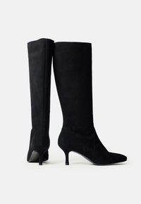 O pereche de cizme negre din suede până la genunchi, cu vârfuri ascuțite și toc subțire de tip kitten, pe un fundal alb.