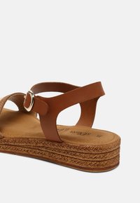 Sandale marron avec une semelle compensée en espadrille tressée, des lanières en cuir lisse et une boucle ronde en accent. Semelle intérieure douce conçue pour le confort.