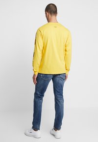 Gul långärmad skjorta i bomull med grafik på ärmarna och bakre logotyp. Bärs med blå jeans och vita sneakers.