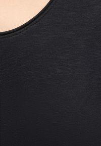 Gros plan sur un tissu noir avec une texture fine en maille et une couture courbée près du coin supérieur, montrant une partie de la peau claire au-dessus du bord.