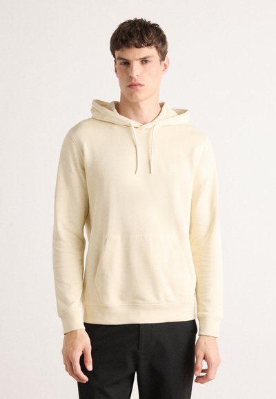 Hoodie couleur crème avec une poche avant, capuche à cordon et poignets côtelés. Texture douce et branding minimal sur la poitrine.