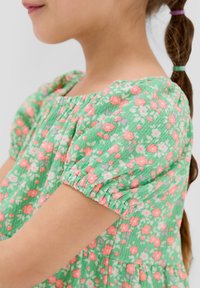 Robe fleurie verte avec des manches courtes bouffantes, ornée de petites fleurs roses et blanches sur un tissu texturé. Une taille froncée apporte du volume.