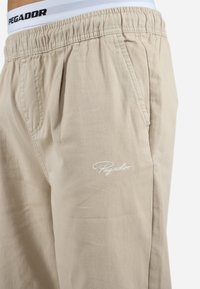 Beige bomull-blandingsbukser med elastisk midje, med hvit logo, sidelommer og en avslappet passform. Jevn tekstur.