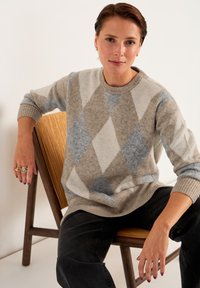 Grå och beige argyle-mönstrad tröja med rund halsringning och ribbade ärmar, bärs med svarta jeans, sittande på en trästol.