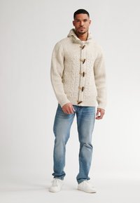 Maglione a coste a treccia color crema con chiusure in legno, indossato sopra jeans blu sbiaditi e sneakers bianche. Texture morbida e vestibilità rilassata.