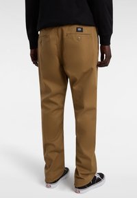 Brązowe bawełniane spodnie o prostym kroju, z dwoma kieszeniami z tyłu zamykanymi na guziki i metką Vans na talii. Na nogach czarne trampki.