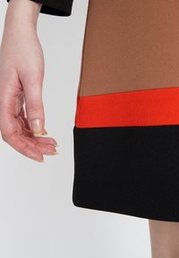 Tailliertes Kleid mit einem zweifarbigen Design aus braunem, orangefarbenem und schwarzem strukturiertem Stoff, mit einer sichtbaren Hand in der Nähe des Saums.