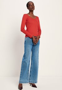 Frau steht mit gefalteten Händen, trägt ein langärmliges rotes Oberteil, helle weite Jeans und dunkle spitze High Heels vor einem schlichten Hintergrund.