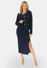 Granatowy garnitur składający się z dopasowanej marynarki z złotymi guzikami oraz pasującej spódnicy ołówkowej, z bocznym rozcięciem i smukłym fasonem.