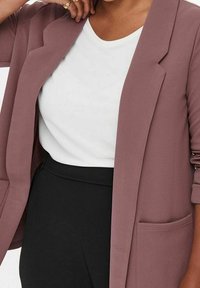 Blazer color malva in tessuto testurizzato, con apertura frontale e due tasche laterali, abbinato a una t-shirt bianca e pantaloni neri.