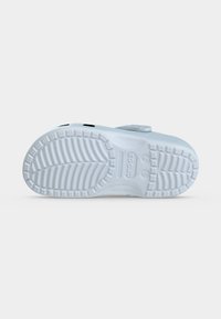 Semelle d'un sabot blanc Crocs montrant un motif profond de la bande de roulement et des indications de taille sur un fond blanc uni.