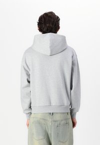 Sweatshirt gris avec capuche, présentant une coupe décontractée et des poignets côtelés. Le tissu semble doux et texturé, avec un design simple et minimal.