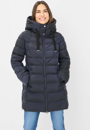 Lächelnde Frau, die eine schwarze, gesteppte Daunenjacke mit hohem Kragen und Kapuze trägt und die Hände in den Taschen hat, vor einem einfarbigen Hintergrund stehend.