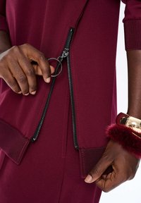 Close-up van donkerhuidige handen die een grote zwarte rits van een bordeauxrode kledingstuk met ribbelmanchetten aanpassen, met een gouden armband met bordeauxkleurige bont.