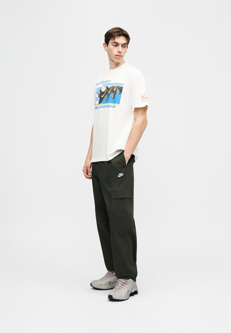 Junger Mann trägt ein weißes T-Shirt mit Grafik, dunkle Nike-Cargohosen und hellgraue Nike-Sneaker, steht mit einer Hand in der Tasche und schaut seitlich.