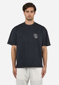 Tricou oversized de culoare albastru închis, cu guler rotund și mâneci scurte, având un logo alb circular pe partea stângă a pieptului.
