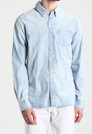 Mand iført en lyseblå denimskjorte med knapper og brystlomme samt hvide bukser foran en ensfarvet baggrund.