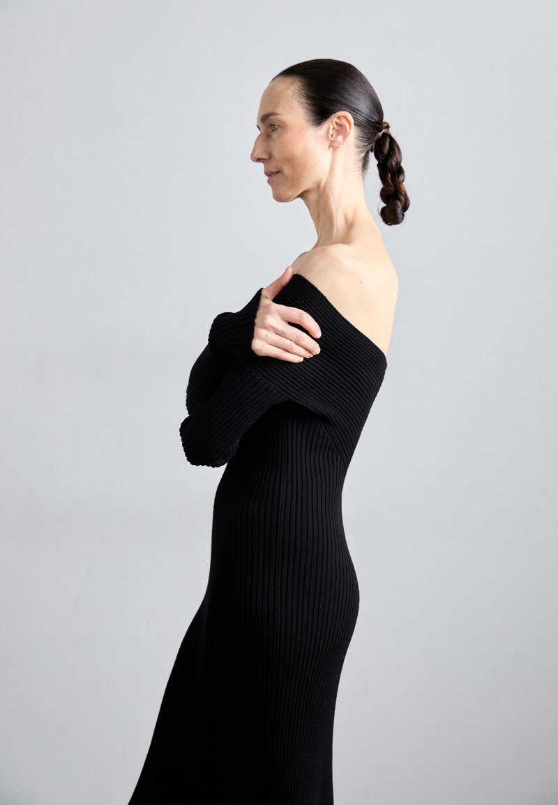 Robe en tricot noir à côtes avec design décolleté, manches longues et silhouette ajustée. Fond gris neutre.