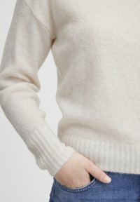 Maglione lavorato a maglia di colore crema con polsini e bordo a coste, caratterizzato da una texture morbida. Mano infilata nella tasca di jeans blu.
