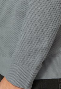 Grau strukturierter Pullover mit einem Waffelstrickdesign, gerippten Bündchen und einem glatten Saum. Der Stoff wirkt weich und atmungsaktiv.