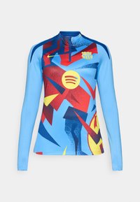 Długorolny top sportowy z żywym niebieskim tłem, zdobiony abstrakcyjnymi, czerwonymi, żółtymi i czarnymi wzorami geometrycznymi. Zawiera logo Nike oraz herb klubu.