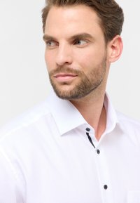 Man met kort bruin haar en een baard, gekleed in een witte, gestructureerde, knoopshirt, kijkt naar links tegen een effen achtergrond.