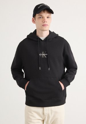 Zwarte hoodie met logo, trekkoorden en een voorzak; gecombineerd met crèmekleurige broek; eenvoudige achtergrond, met een mannelijke model.