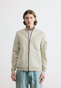 Νέος άνδρας με μεσαίου μήκους καστανά μαλλιά, φοράει μπεζ ελαφρύ μπουφάν με φερμουάρ και ανοιχτό μπλε τζιν, στέκεται μπροστά σε λευκό φόντο.