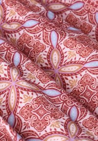 Stoff mit einem sich wiederholenden floralen und geometrischen Muster in Rot, Blau und Gold auf einem hellrosa Hintergrund, mit filigranen Details.