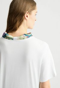 Maglietta bianca a maniche corte, con un colletto decorato da un motivo colorato. Il colletto presenta disegni floreali e geometrici nei colori blu, rosso e verde.