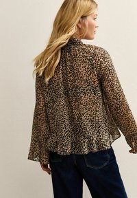 Vrouw met lang blond haar, gekleed in een blouse met een bruin en zwart luipaardpatroon en donkerblauwe spijkerbroek, met de rug naar de camera toe tegen een plain muur.