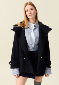 Cappotto peacoat nero con cappuccio, bottoni dorati e tasche laterali, indossato sopra una camicia a strisce blu e bianche, abbinato a una gonna blu navy.
