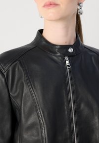 Črna usnjena jakna z visokim ovratnikom, srebrnim zadrgo in subtilnimi detajli šivanja. Gladka tekstura z oprijetim dizajnom.