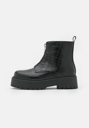 Botte noire à la cheville avec texture crocodile, fermeture éclair à l'avant, semelle épaisse à crampons et languette de traction sur un fond blanc uni.