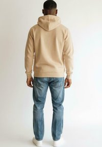 Homme portant un hoodie beige, un jean bleu et des baskets blanches, se tenant de dos contre un fond clair uni.