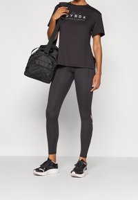 Person som bär en svart Hyrox fitness t-shirt, svarta leggings med sidologga, svarta sneakers och har en svart träningsväska över axeln.