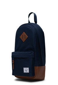 Mochila de color azul marino con un fondo de cuero marrón, parche en forma de diamante, bolsillo frontal con cremallera y etiqueta de logo. Fabricada con tejido resistente.