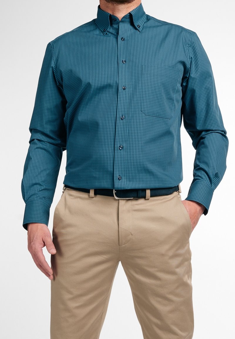 Turquoise overhemd met een klein geruit patroon, lange mouwen en een borstzakje. Gecombineerd met lichtbeige broek en een zwarte riem.