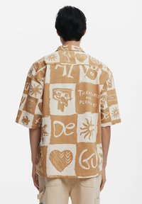 Kortærmet skjorte i beige og hvid med grafiske tryk; har en boxy pasform, krave og forskellige håndtegnede mønstre.
