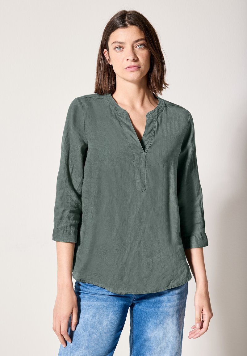Blusa verde, leggera, a maniche lunghe con scollo a V e una vestibilità rilassata, con una finitura testurizzata e un orlo arrotondato. Abbinata a jeans blu.